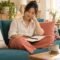 Woman looking at an open notebook while sitting on a teal sofa in a sunlit room.