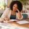 Woman writing in a notebook beside a paper calendar at a sunlit kitchen table.