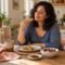 Woman enjoying toast at a sunlit kitchen table with yogurt, berries, and chocolate nearby.